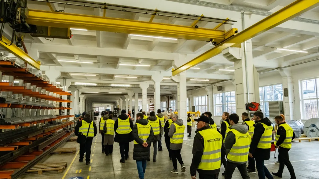 Grupul de vizitatori traversând hala de producție pe sub podul rulant